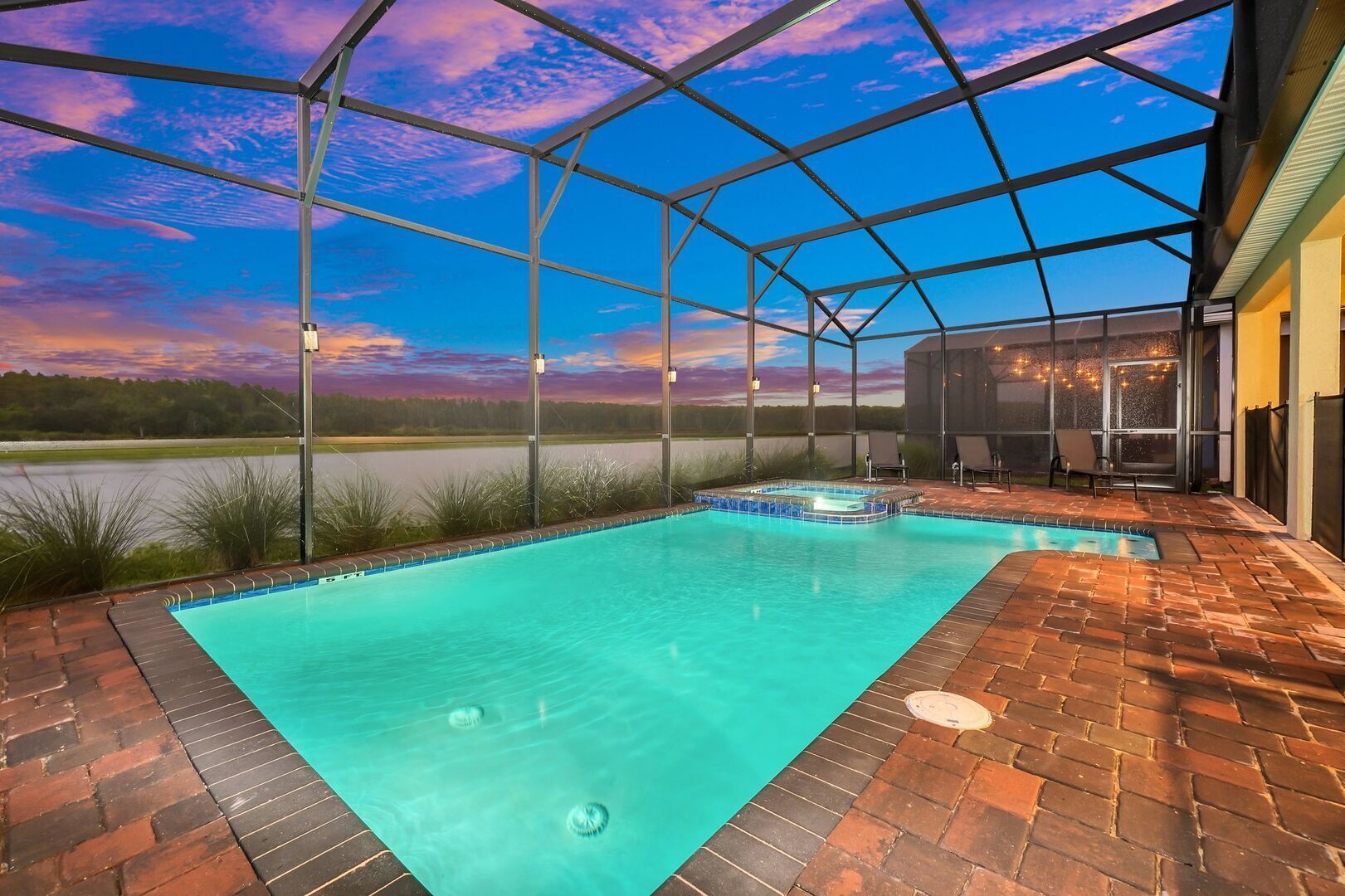 A swimming pool with a brick patio and a covered area with chairs and a grill, overlooking a body of water.