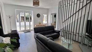 A modern living room with a black leather sofa, a glass coffee table, and a staircase with a black railing. There is a large window with a view of the outside, and a clock on the wall.