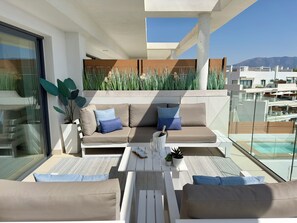 A modern outdoor seating area with a sofa and chairs, a small table with a bottle and glasses, and a view of a pool and buildings. - OneHeights: Luxuriously equipped apartment in prime location with phenomenal view! (La Cala de Mijas)