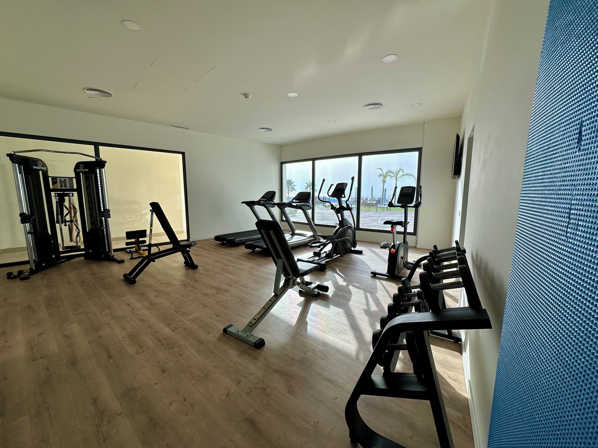 Une salle de sport équipée de divers appareils d’exercice, dotée de grandes fenêtres et d’un plancher en bois.