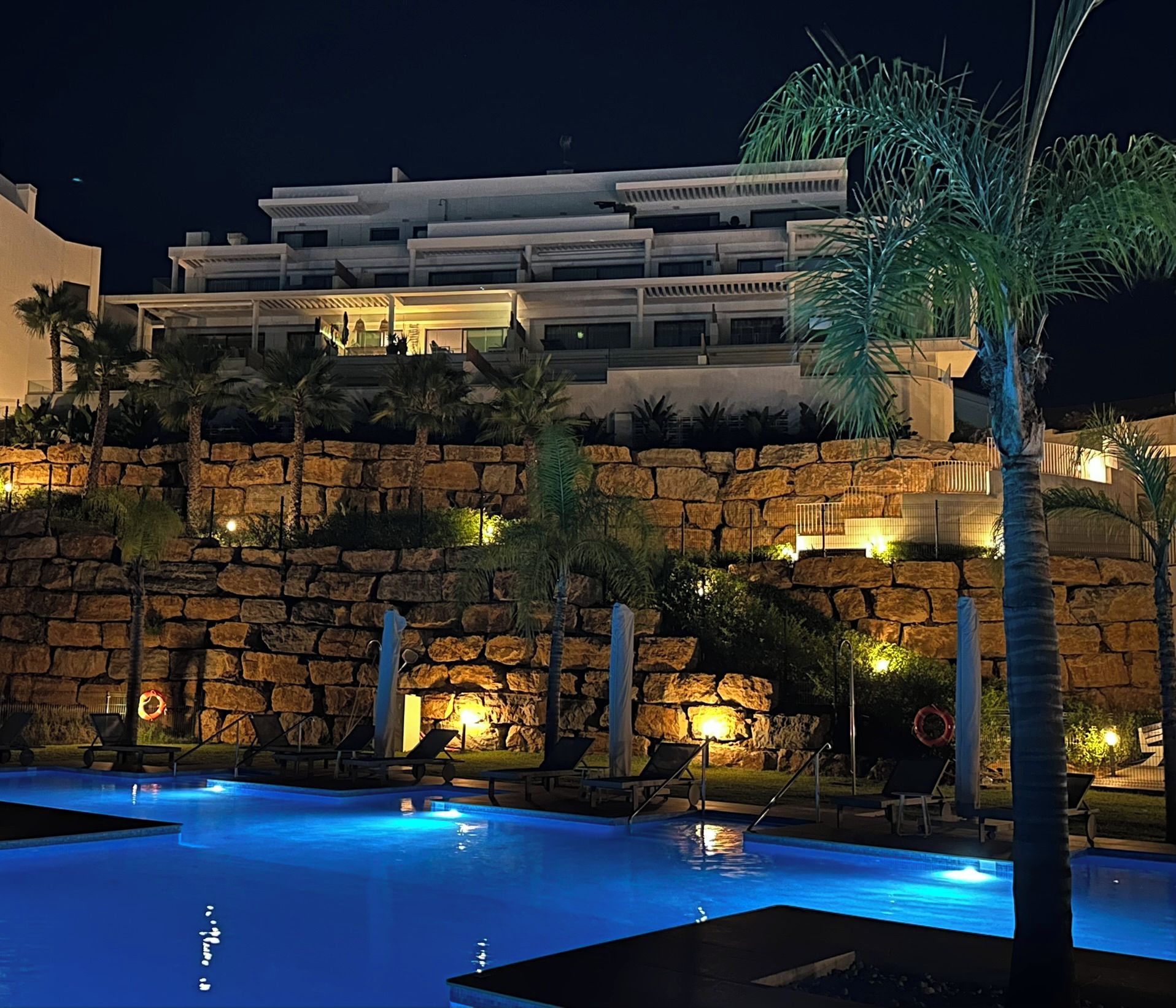 A nighttime view of a hotel with a swimming pool, stone walls, and palm trees.