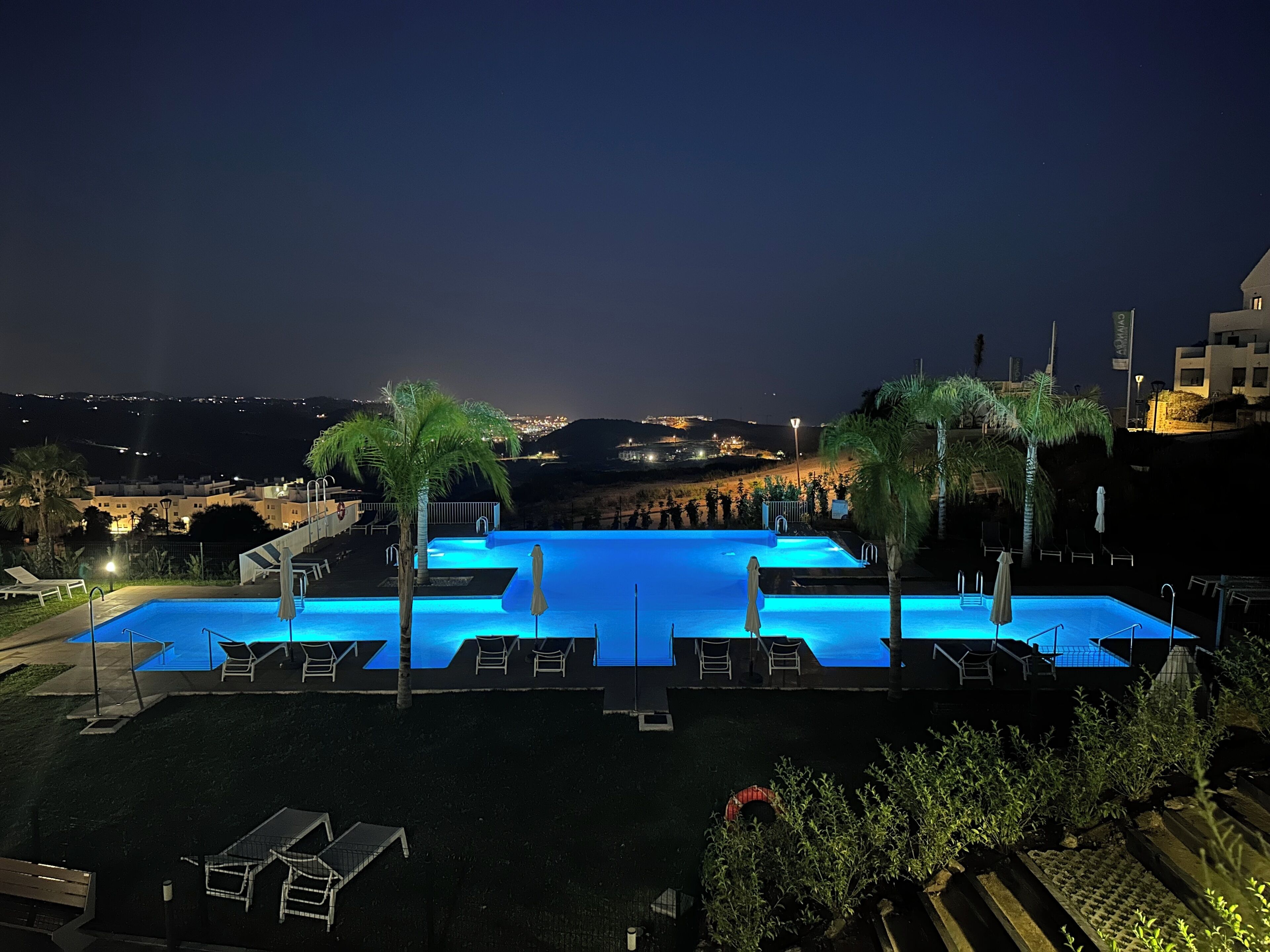 A nighttime view of a rectangular swimming pool with blue lighting, surrounded by lounge chairs and palm trees, with a cityscape in the background.