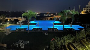 Une vue nocturne d’une piscine rectangulaire illuminée de lumières bleues, entourée de chaises longues et de palmiers, avec un paysage urbain en arrière-plan.
