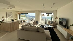 Un salon moderne avec un canapé, une table basse et un téléviseur, de grandes fenêtres offrant une vue sur la ville, et un coin repas avec une table et des chaises.