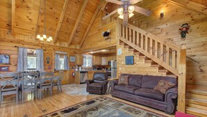 Interior espacioso de una cabaña de madera con comedor, sala de estar con sofá y sillones, cocina y una escalera.