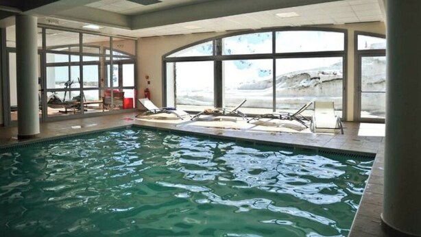 An indoor swimming pool with clear glass walls, lounge chairs, and a view of the outdoors. - T2 swimming pool, terrace, private parking R. Pic du Midi (Bagnères-de-Bigorre)