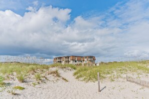 A beachfront building with a sandy path leading to it, surrounded by grass and dunes.