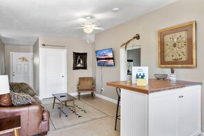 A living room with a brown leather sofa, a wooden chair, a small table, a ceiling fan, a TV, and a kitchen area with a countertop and cabinets.
