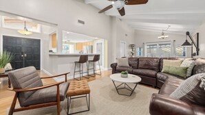 Un salon spacieux avec un canapé en cuir marron, un fauteuil en bois et une table basse ronde. On peut voir un ventilateur de plafond et des lampes suspendues.