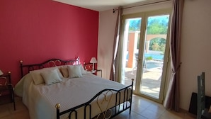 Une chambre à coucher avec un lit, deux tables de chevet avec des lampes, un mur rouge et une porte-fenêtre coulissante donnant sur un espace extérieur.