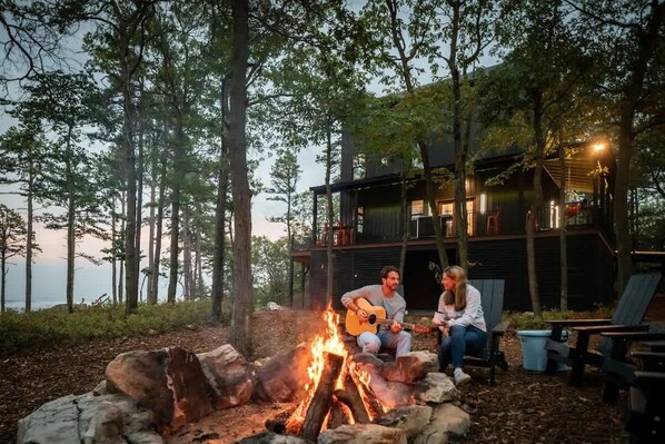 A cabin with a fire pit, surrounded by trees, and a person playing guitar.