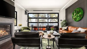 A modern living room with a fireplace, leather sofas, a coffee table, and a large window overlooking a forested area. - Monte Vista~Golf~Views~PS5~Sport Court~EV Charger (Berkeley Springs)