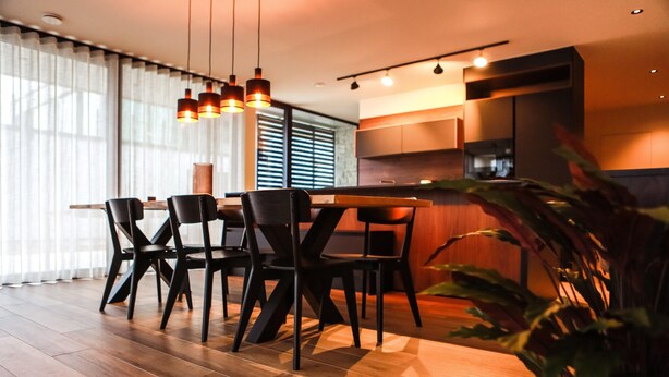 Zona pranzo moderna con tavolo e sedie in legno, cucina sullo sfondo e un'ampia finestra con tende.