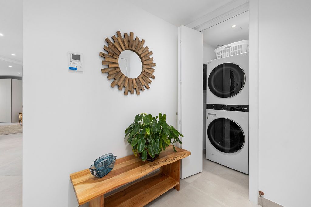 A modern laundry room with two stacked washing machines, a wooden bench with a potted plant, and a wall-mounted mirror.