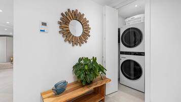 A modern laundry room with two stacked washing machines, a wooden bench with a potted plant, and a wall-mounted mirror.