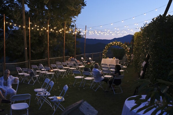 An outdoor event space with rows of chairs and tables, string lights overhead, and a backdrop of trees and mountains.