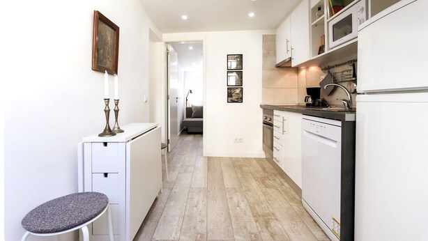 Una cocina estrecha con armarios blancos, una encimera y un lavavajillas. Hay un taburete pequeño y un cuadro en la pared.