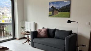 A modern living room with a grey sofa, a wooden ceiling, a large window with a view of mountains, and a painting of a mountain landscape. - Orchard Villa - Loft Apartment (Bled)
