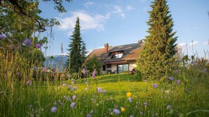 A house with a sloped roof, surrounded by tall trees and a grassy field with wildflowers. - Orchard Villa - Loft Apartment (Bled)
