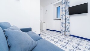 A room with a blue sofa, a flat-screen TV mounted on the wall, a window with blue patterned curtains, and a tiled floor with a blue border. - Van Gogh Room (Amalfi)