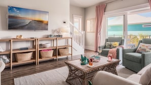 Un salon spacieux avec une grande télévision, des meubles en osier et une vue sur une maison, visible à travers une porte-fenêtre coulissante.