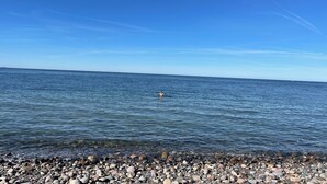 Ein freier Blick auf einen Kiesstrand, der zu ruhigem Wasser führt, mit einer Person, die in der Ferne schwimmt.