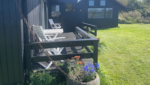 Eine Holzterrasse mit zwei Liegestühlen und einer Bank, umgeben von einem blühenden Garten und Blick auf ein Haus.