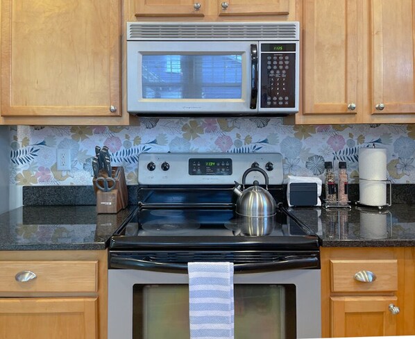 A kitchen with wooden cabinets, a stainless steel microwave, a stove, and a kettle.