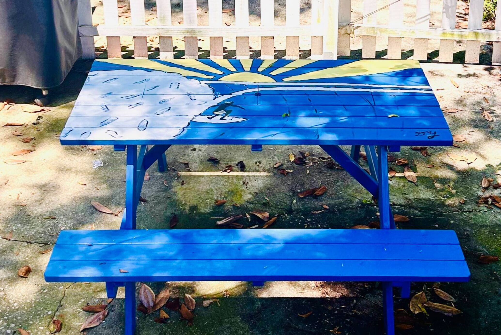 Une table de pique-nique bleue, décorée d’un motif peint représentant un soleil et des nuages, entourée d’une clôture blanche en piquets.