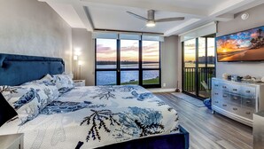 Ein Schlafzimmer mit einem großen Bett, einem Deckenventilator, Blick auf den Sonnenuntergang durch das Fenster und einem Fernseher an der Wand.