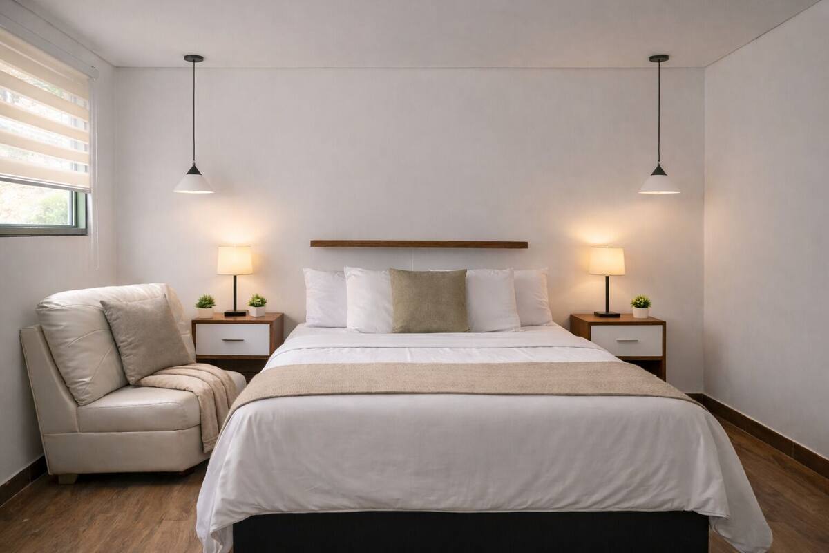 Habitación de hotel moderna con una cama grande, dos mesitas de noche con lámparas, una silla y una ventana con persianas.