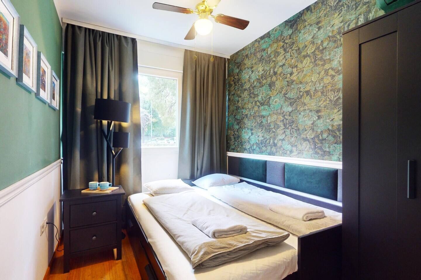 Une chambre à coucher comprenant un lit, une table de chevet, une lampe, un ventilateur de plafond et une fenêtre avec des rideaux.