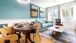 A dining area with a round table, chairs, and a sofa. There are two paintings on the wall and a window with curtains. - Gallery Gardens Apartment (Split)