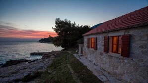 A stone building with a red-tiled roof, located by a rocky shoreline with a view of the sea and a sunset in the background.