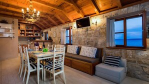 A cozy room with a stone wall, wooden ceiling, a dining table with chairs, a sofa with cushions, and a television mounted on the wall.
