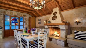 A rustic interior with a stone fireplace, wooden ceiling beams, and a dining area with white chairs.