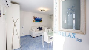 A modern living room with a white sofa, a glass dining table, and a painting of a coastal scene. - De Vivo Realty - Blue Angel Relais (Meta)