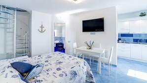 A modern hotel room with a bed, a desk, a TV, a bathroom, and a kitchen area. - De Vivo Realty - Blue Angel Relais (Meta)