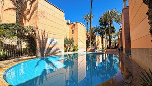 A swimming pool surrounded by buildings with palm trees, a clear blue sky, and a sign indicating a children's play area. - 10 minutes from the Majorelle garden Riad Beldi Chic An Authentic Stay”- Abritel (Marrakech)