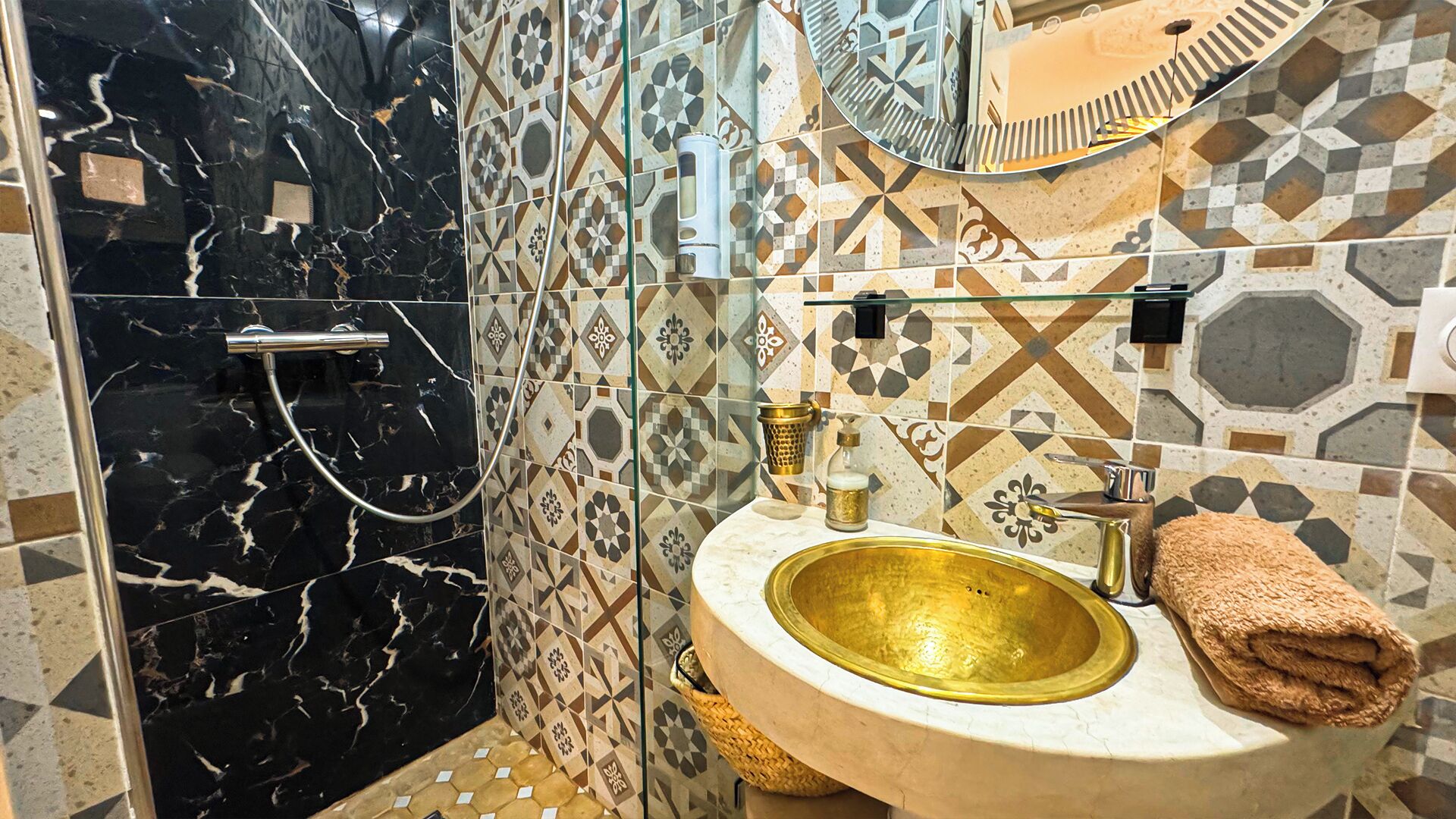 Une salle de bain dotée d’une douche en marbre noir, d’un lavabo avec un receveur doré et d’un miroir agrémenté d’un pare-splash en carreaux géométriques.