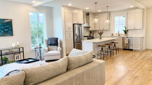 A modern kitchen with white cabinetry, a central island with stools, and a dining area with a sofa and armchair. - Newly built 4BR, close to Duke, ready for Duke Graduation! (Durham)