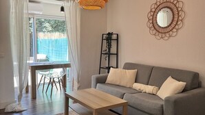 Een woonkamer met een grijze bank, een houten salontafel, een geweven vloerkleed, een hanglamp, een spiegel, en een eethoek met een tafel en stoelen.