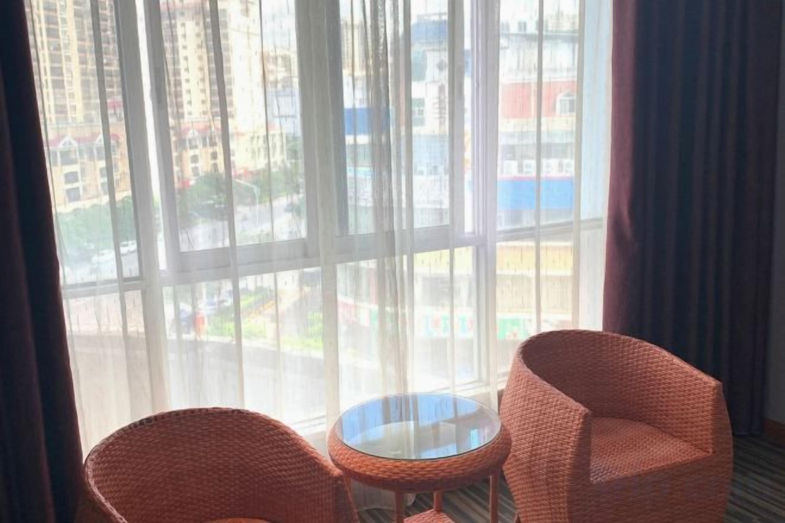 Una habitación con dos sillones de mimbre y una mesita, ventanas amplias con vistas a edificios y una carretera.