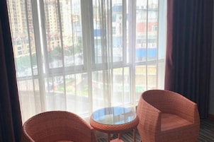 Una habitación con dos sillones de mimbre y una mesita, ventanas amplias con vistas a edificios y una carretera.