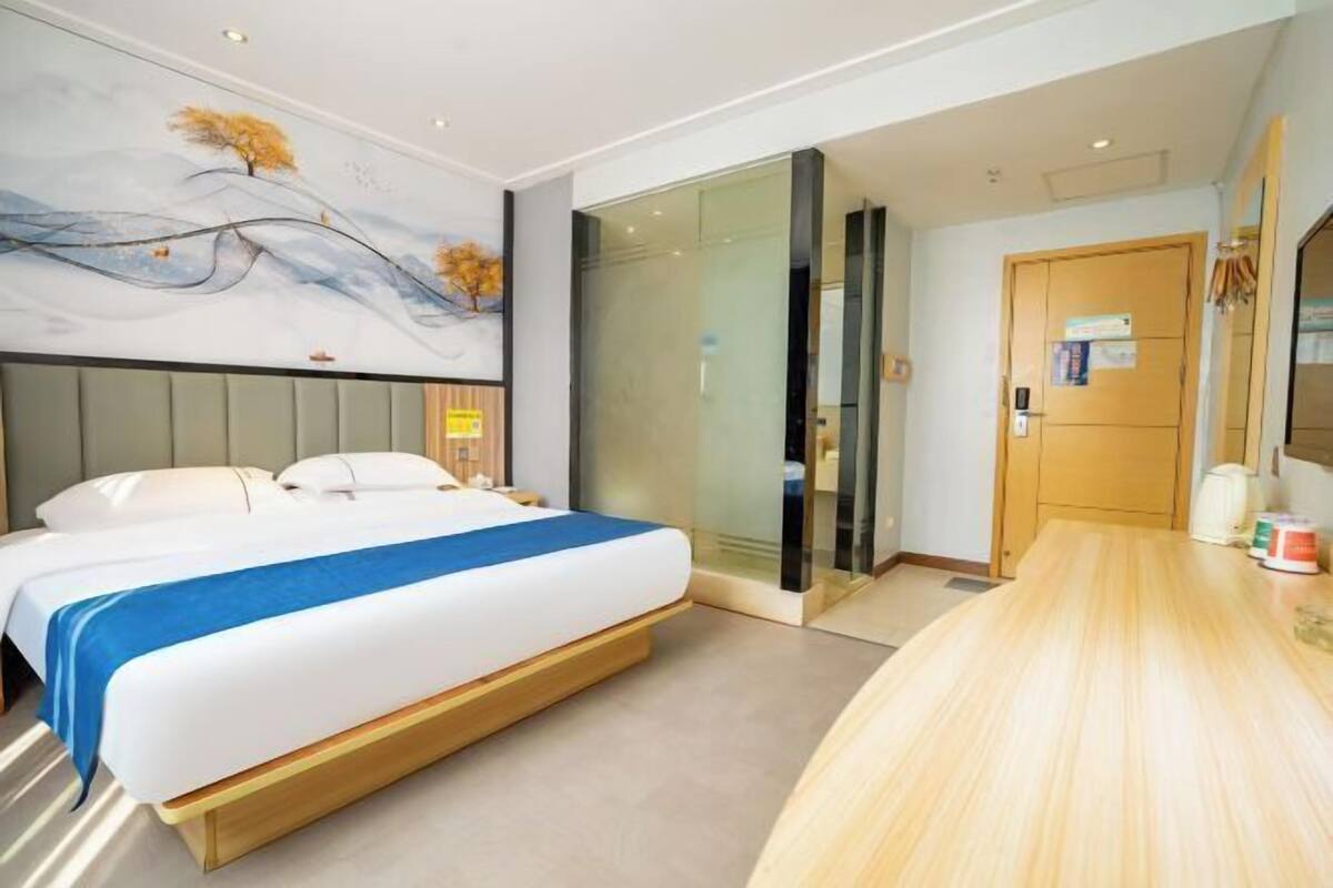 Une chambre d’hôtel moderne avec un grand lit, une cloison en verre, un bureau en bois et un tableau au mur.