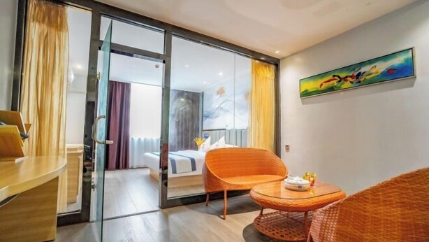 Quarto de hotel com porta de vidro para uma varanda, cama com cabeceira branca, uma cadeira laranja, uma mesinha redonda e um quadro na parede.