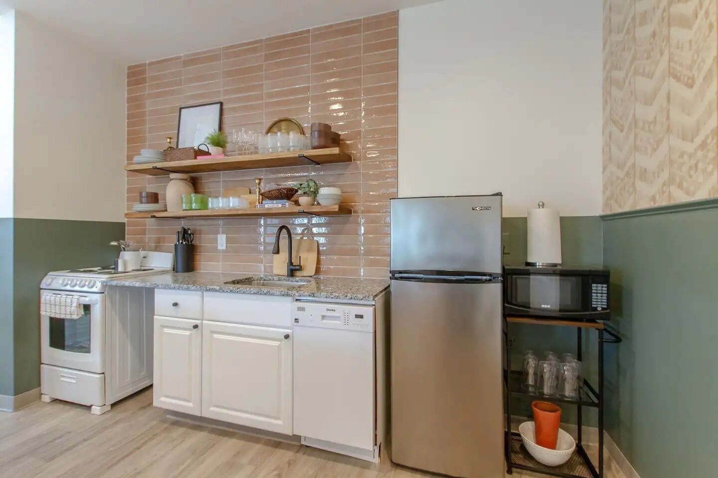 A compact kitchen with a stainless steel refrigerator, a microwave, a dishwasher, and a stove. There are shelves above the sink holding various kitchen items.
