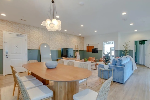 A modern living room with a wooden dining table, white cushioned chairs, a blue sofa, and a hanging light fixture.