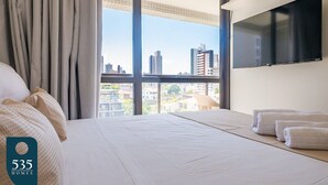 Hotellihuone, jossa on suuri ikkuna, valkoisilla lakanoilla varustettu sänky ja seinälle asennettu taulutelevisio.