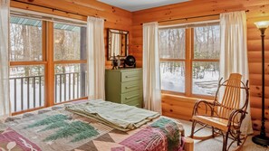 Un dormitorio de cabaña de madera con cama, cómoda, mecedora y ventanas amplias.